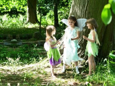 Children dressed as fairies playing in lush Capel Manor Gardens, surrounded by trees and greenery.