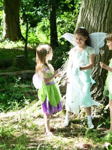 Children dressed as fairies playing in lush Capel Manor Gardens, surrounded by trees and greenery.