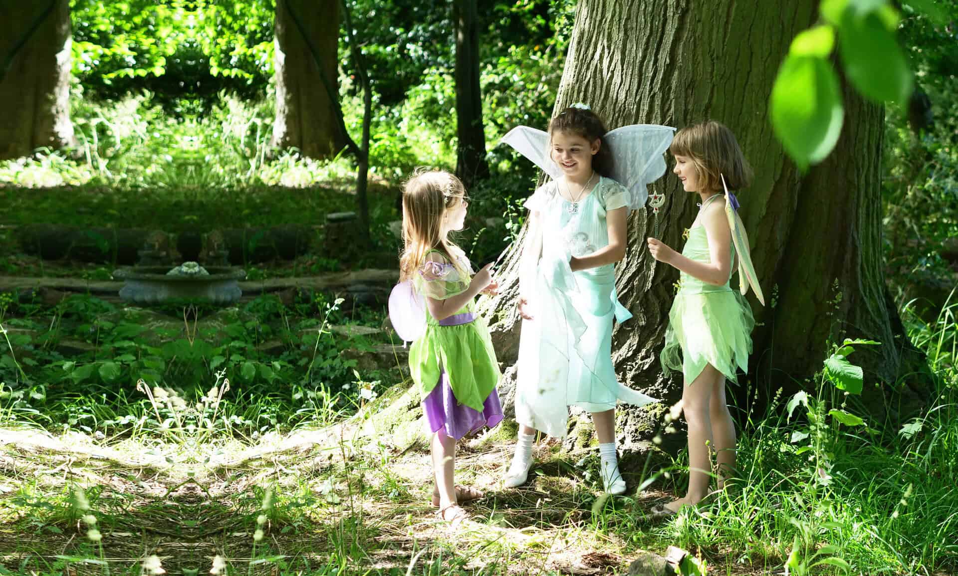 Children dressed as fairies playing in lush Capel Manor Gardens, surrounded by trees and greenery.